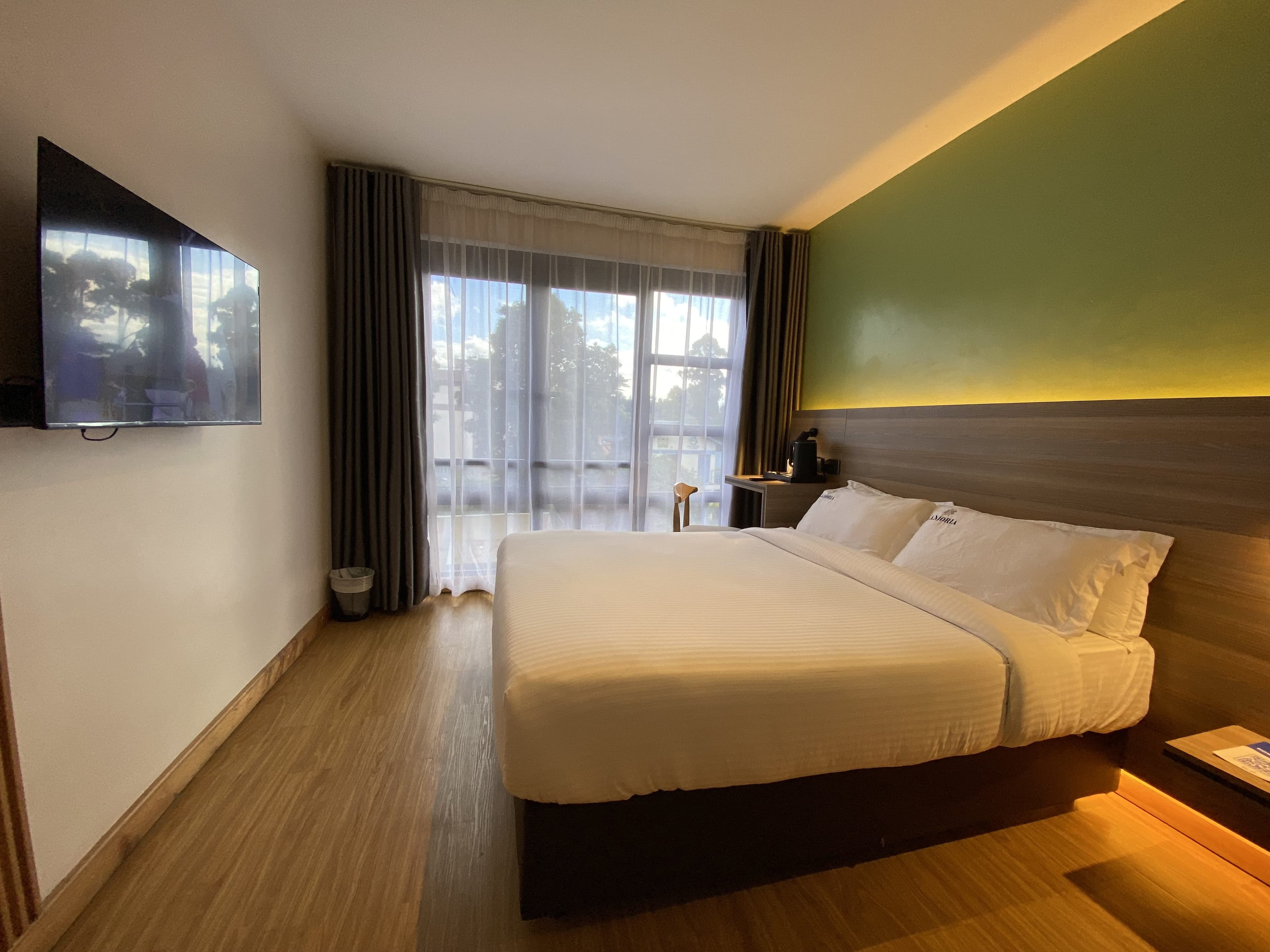 Sunlit guest room with a made white bed, warm under-lit green feature wall and trees through the window